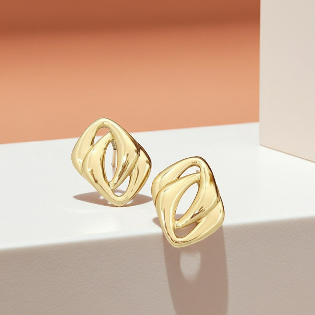 A pair of gold-toned geometric stud earrings with an abstract, interlocking loop design, displayed on a white platform against a peach and cream background.