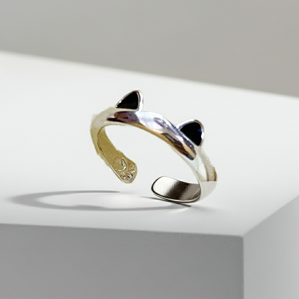 A silver-toned open cuff ring designed like a cat's head, featuring pointed ears with black enamel details and a polished band, displayed on a white geometric pedestal.