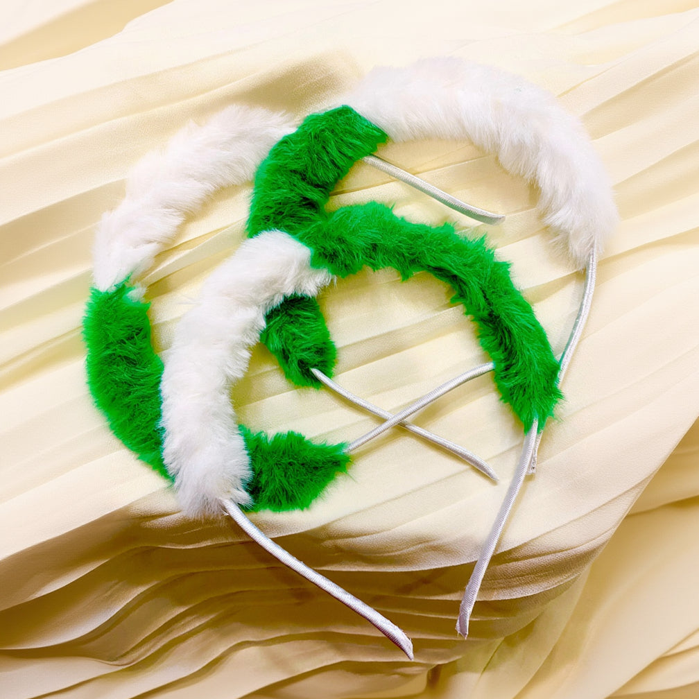 A green and white furry headband with thin silver metal wire arms, laid flat on a cream-colored pleated fabric background.