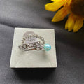 Silver ring with a turquoise bead on a gray display card, next to a yellow flower.