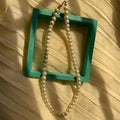 Pearl necklace draped over a teal square frame on a textured beige background