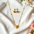 Gold heart-shaped necklace and earrings on a white surface with flowers in the background