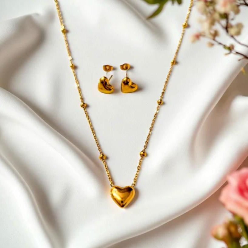 Gold heart-shaped necklace and earrings on a white surface with flowers in the background