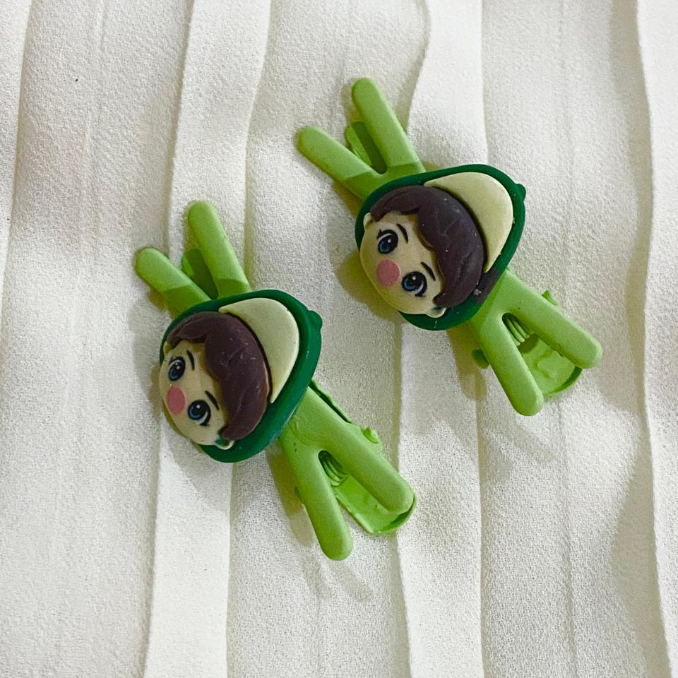 Two green hair clips with cartoon character faces on a white fabric background