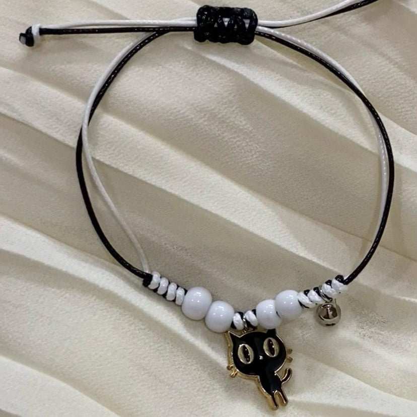 Black cord bracelet with white beads and a cat-shaped charm on a beige fabric background