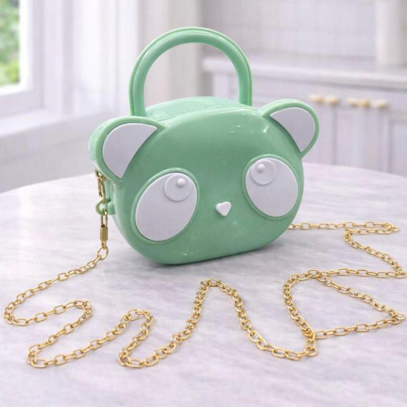 A mint green handbag shaped like a cartoon bear face with white ears and eyes, featuring a gold chain strap, displayed on a light marble table in a bright room.