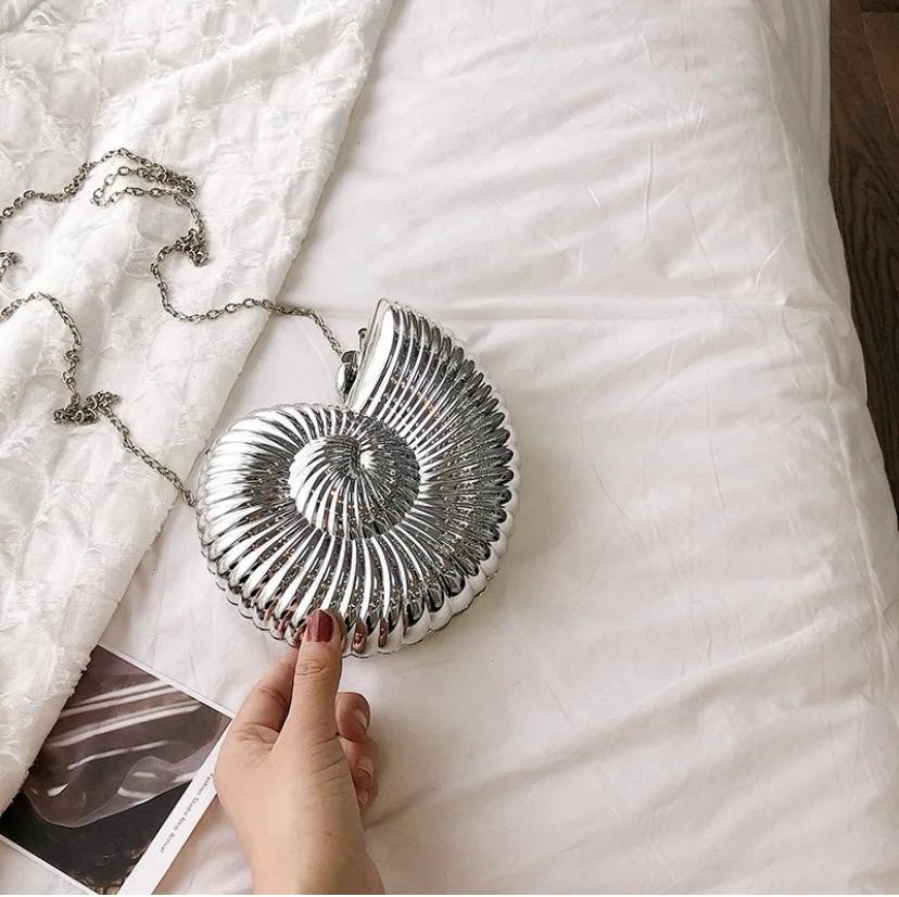 A silver metallic nautilus shell-shaped shoulder bag with a chain strap, held by a hand over a white textured blanket and white bedsheet background.