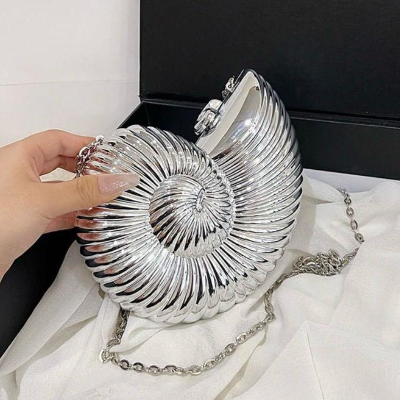 A hand holds a metallic silver shell-shaped shoulder bag with a chain strap, resting on white fabric in front of a black box.