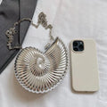 A silver metallic nautilus shell clutch bag with a chain strap, displayed next to a cream-colored smartphone case on a white fabric background.