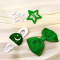 Three hair accessories arranged on a cream pleated fabric background: a white star clip, a green star clip, and a green ribbon bow with a green-and-white crescent-and-star emblem on the clasp.
