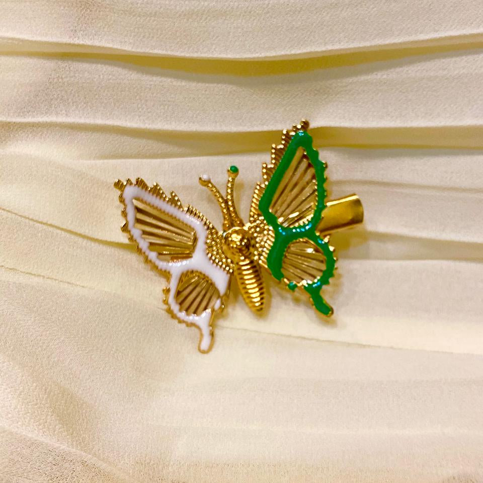 A gold butterfly-shaped hair claw clip with one white wing and one green wing, displayed on a cream-colored pleated fabric background.