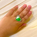 A heart-shaped ring with a green and white crescent moon design set in silver, worn on a child's hand against a cream-colored pleated fabric background.