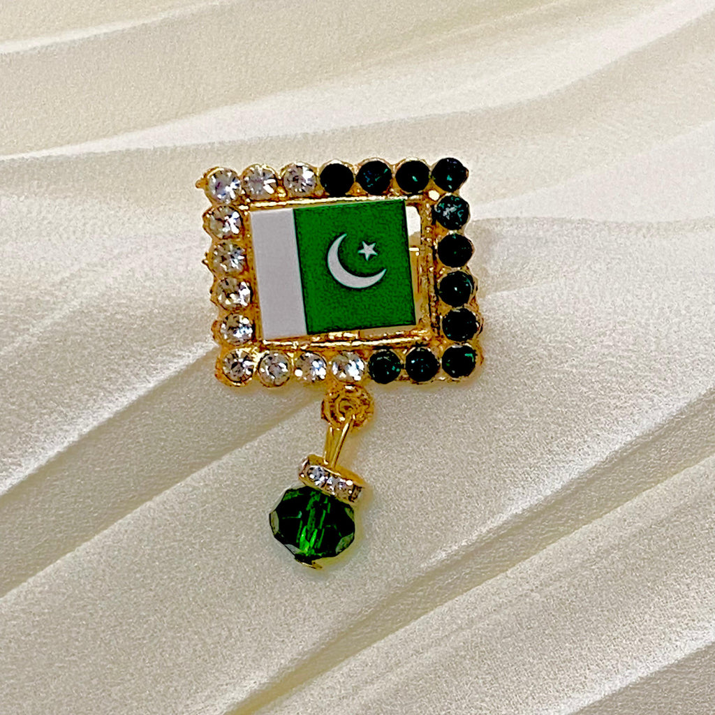 A gold-tone brooch featuring the Pakistan flag with a dangling green faceted crystal, displayed on a cream-colored satin fabric background.