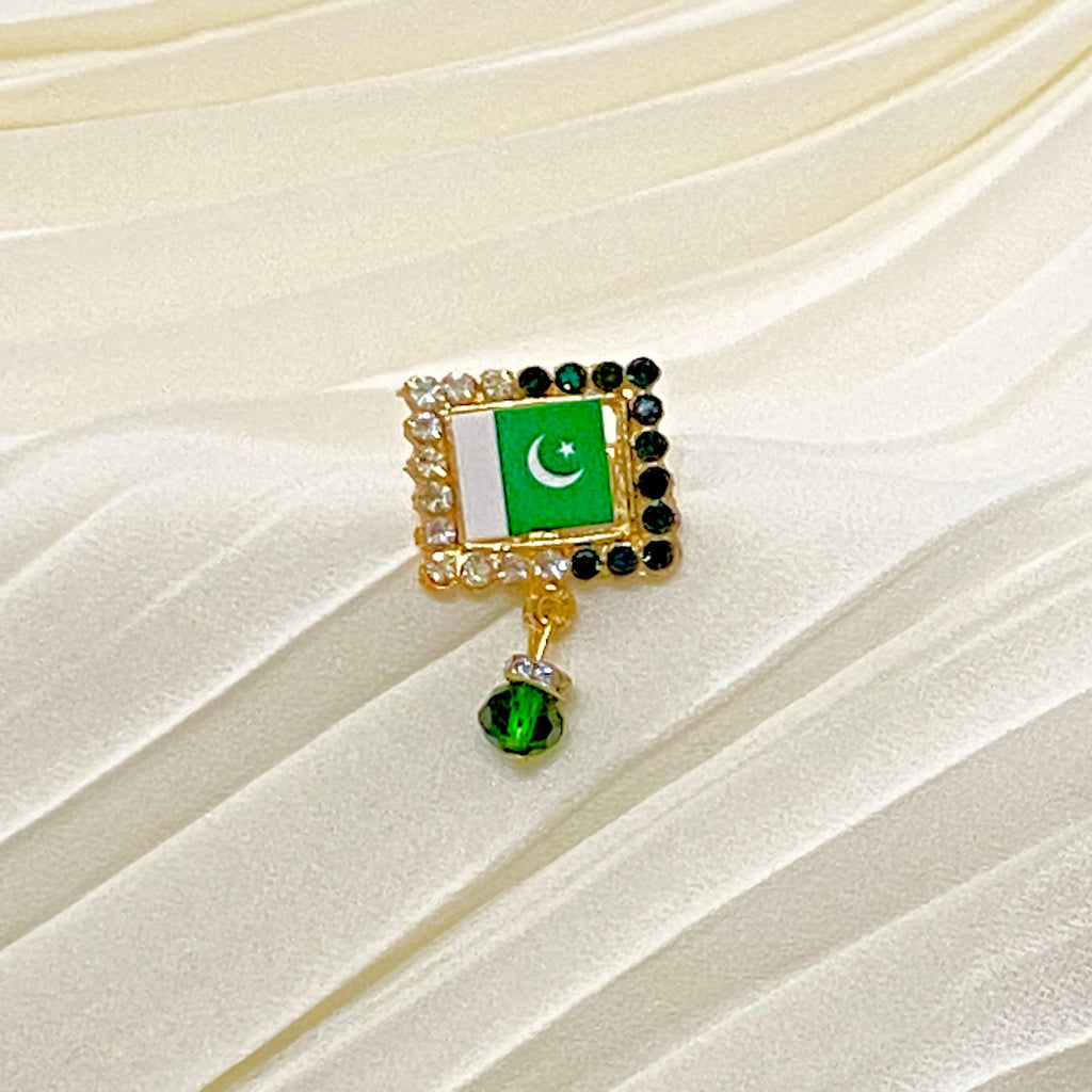 A gold-tone brooch featuring the Pakistan flag with a dangling green crystal charm, displayed on a cream-colored pleated fabric background.