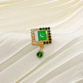 A gold-tone brooch featuring the Pakistan flag with a dangling green crystal charm, displayed on a cream-colored pleated fabric background.