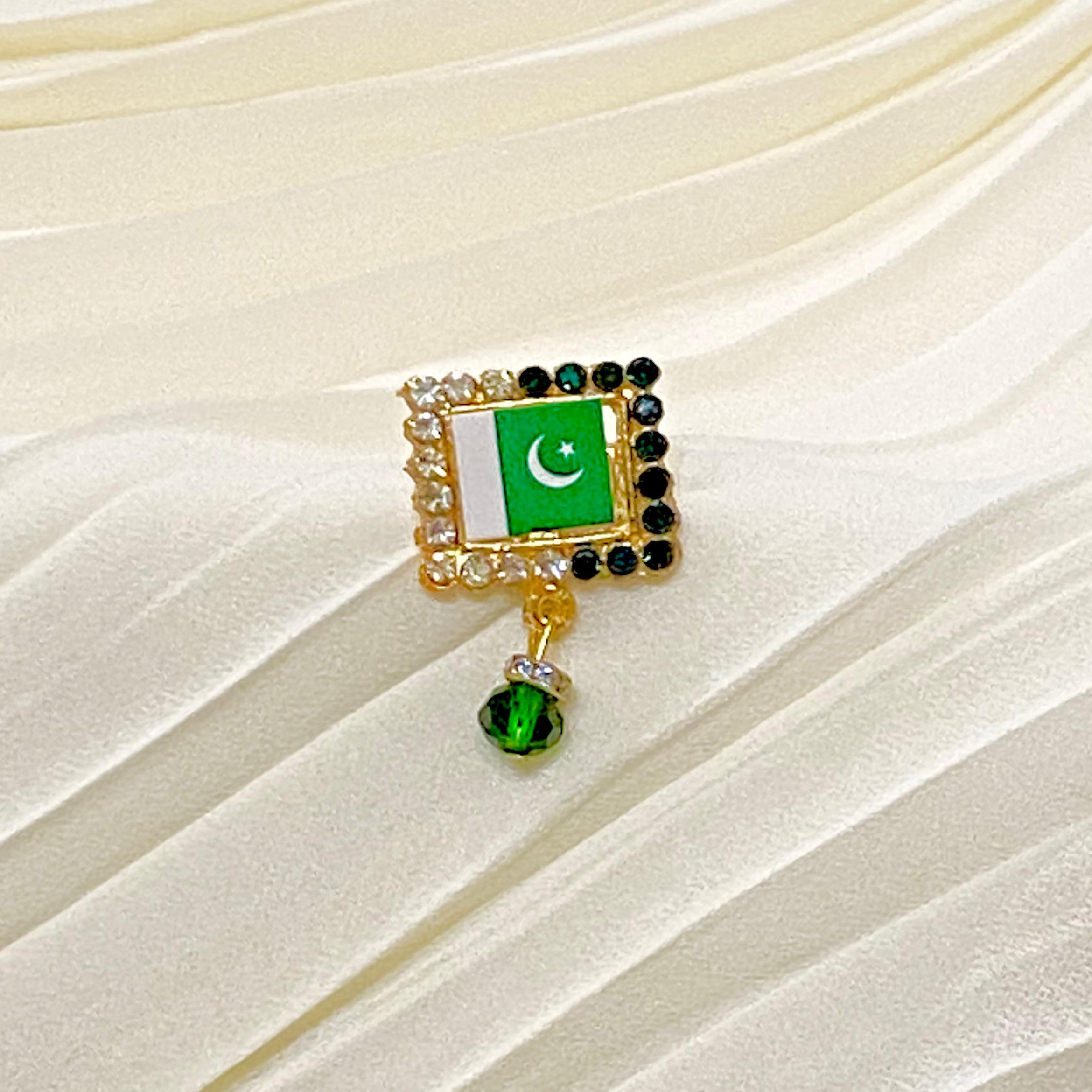 A gold-tone brooch featuring the Pakistan flag with a dangling green crystal charm, displayed on a cream-colored pleated fabric background.