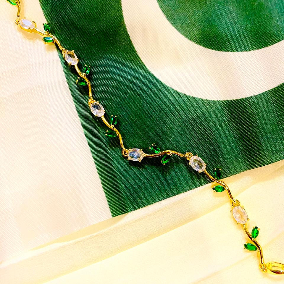 A gold vine-style necklace with green leaf accents and clear oval gemstones, draped diagonally across a green and white flag on a cream fabric background.