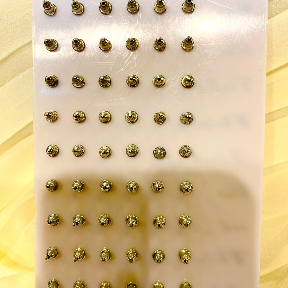 A white rectangular display board holding dozens of small metallic gold oval studs or hardware pieces arranged in rows, photographed against a pale yellow background.