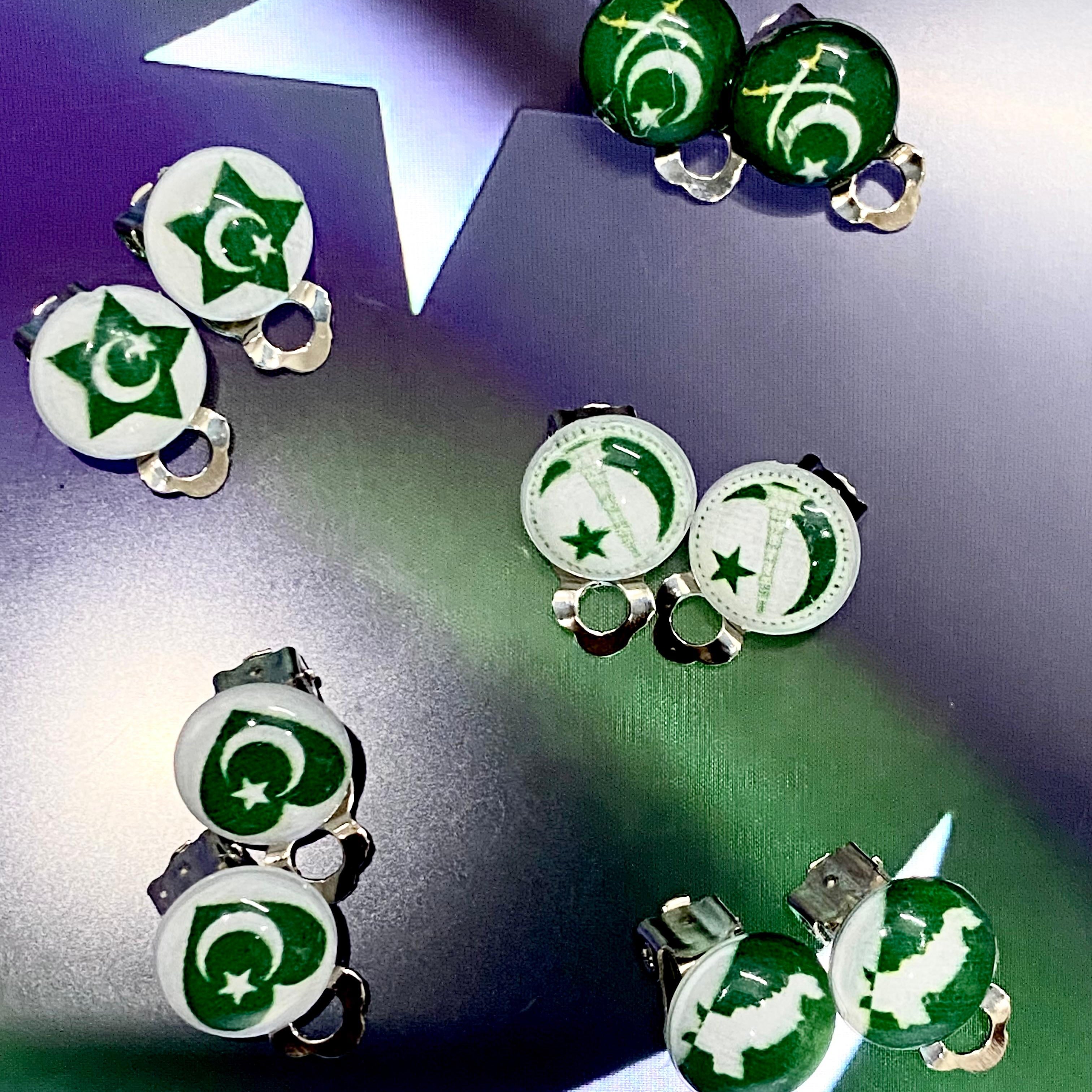 Six pairs of Pakistan-themed stud earrings displayed on a reflective surface with a star-shaped prop. The earrings feature green and white enamel designs with the Pakistani flag stars and crescents, set in silver-tone metal.