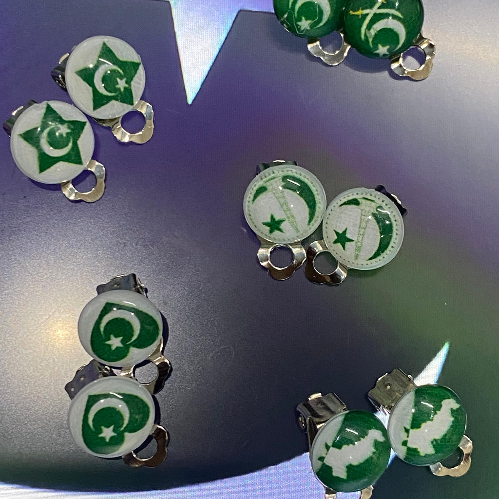 Six pairs of stud earrings displayed on a dark reflective surface with a star-shaped light reflection. The earrings feature green and white enamel discs printed with the Pakistan flag design, attached to silver-toned metal posts.