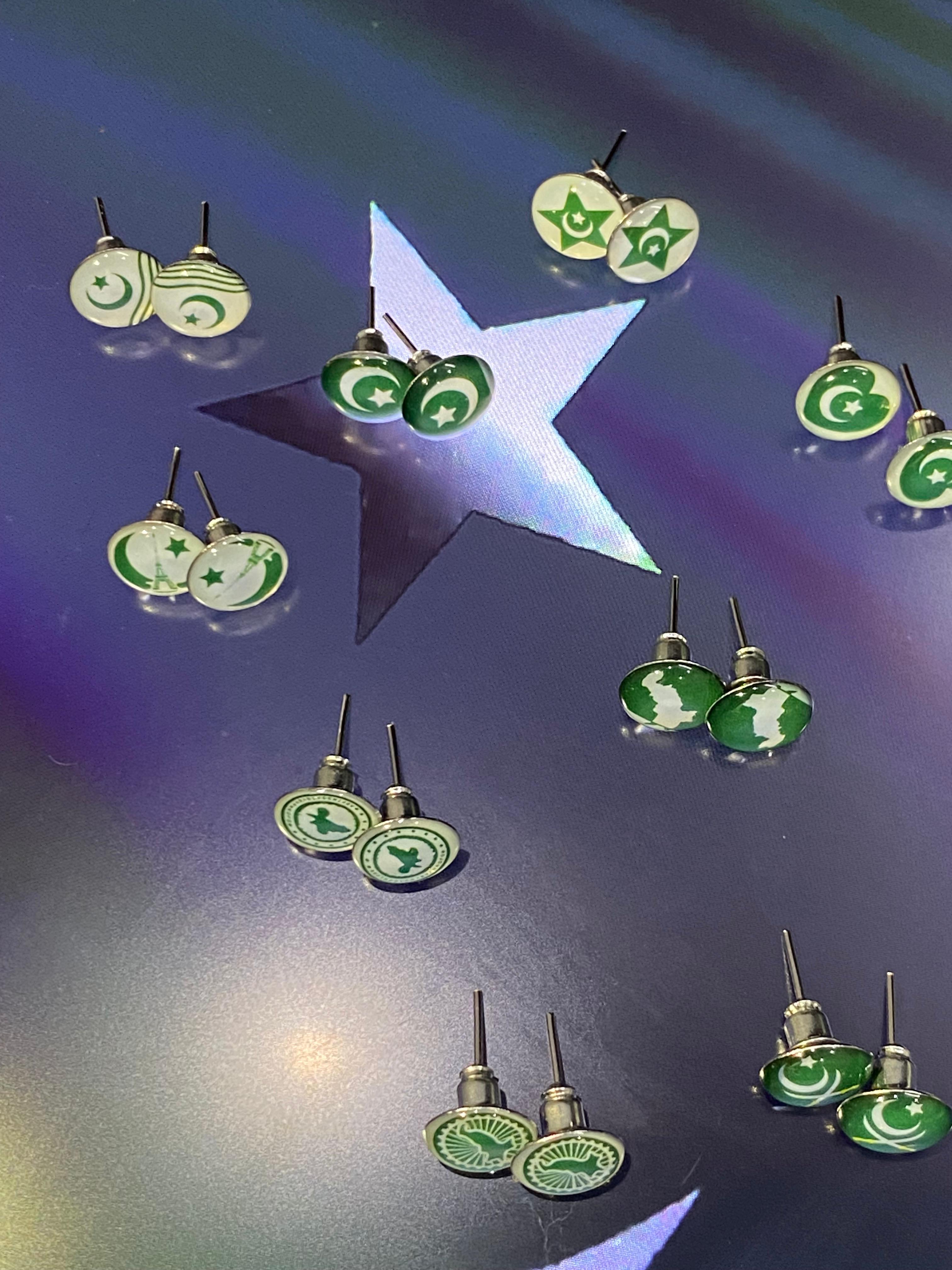 Multiple pairs of green and white enamel stud earrings arranged on a reflective iridescent surface with a large star-shaped cutout in the center.