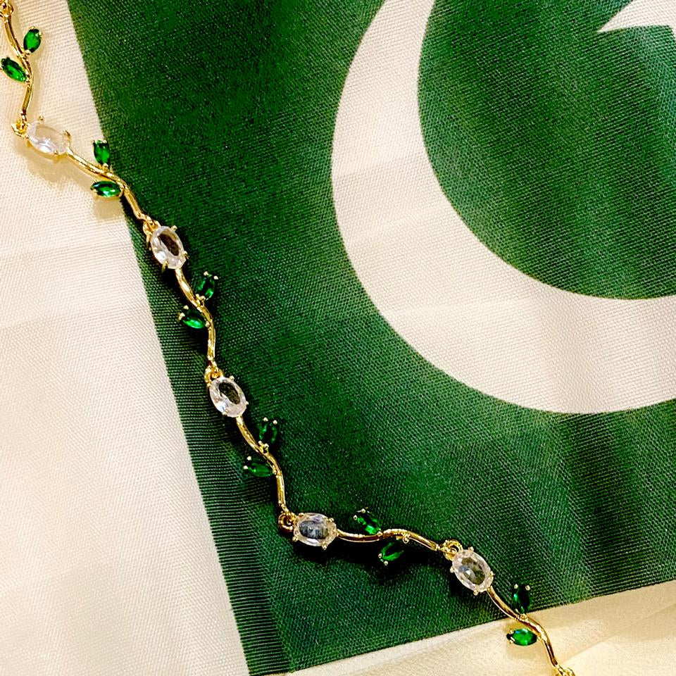A gold vine-style necklace with clear oval crystals and small green leaf accents, draped diagonally over a green and white fabric background.