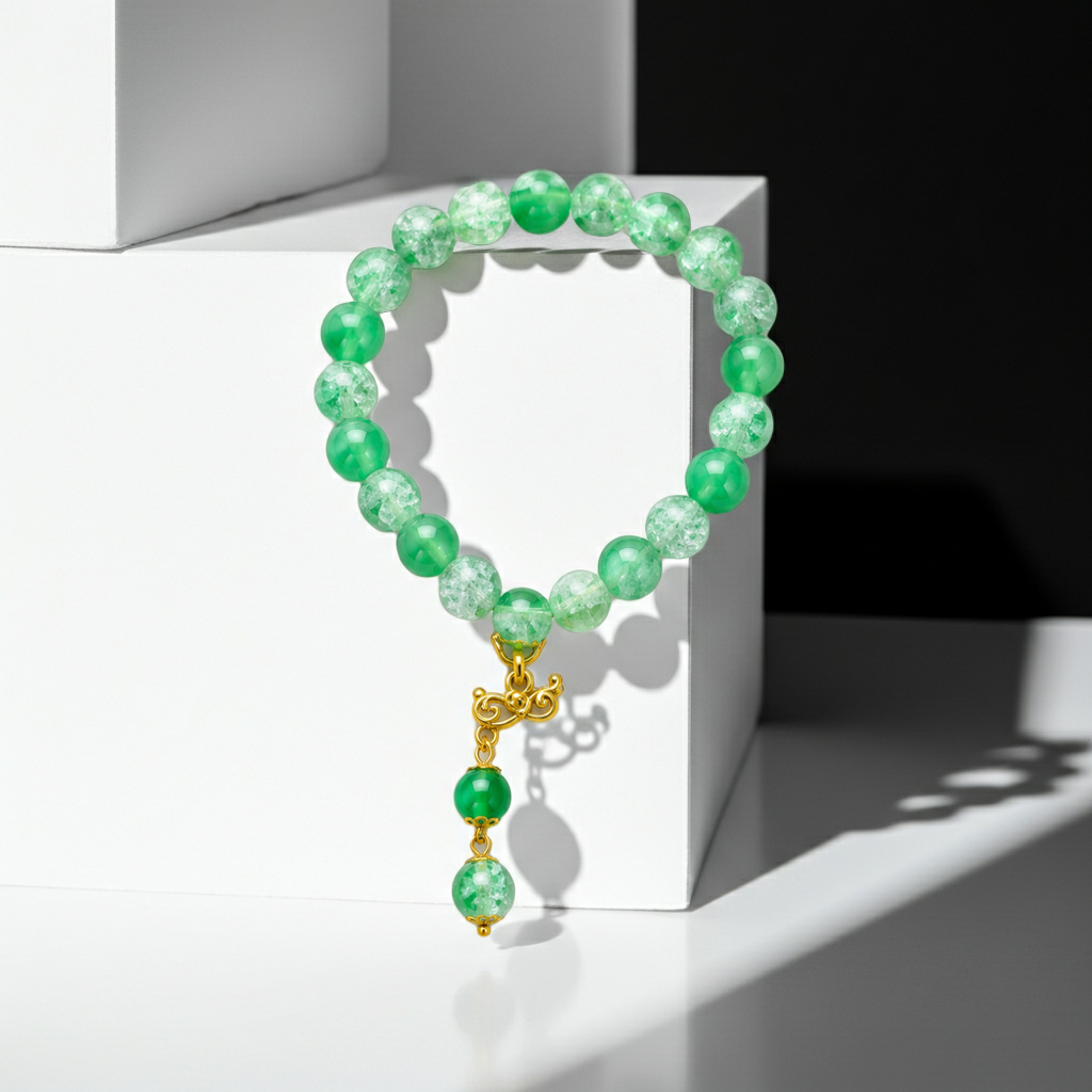 A beaded bracelet made of green marbled and opaque beads with a gold-toned decorative clasp and dangling beads, displayed on a white geometric pedestal.